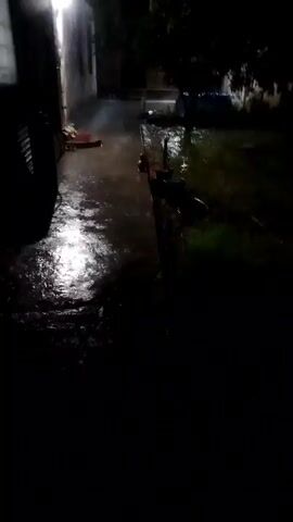 A camera is angled downwards at a dark, flooded courtyard at night. The sound of heavy rain and wind can be heard. A bright light emanates from the left side of the frame, illuminating a wet, concrete path and puddles. In the background, partially obscured by darkness, are trees and what appears to be a blue tarp covering an object. The water is murky and disturbed by the rain. The video is very dark and blurry, making it difficult to discern specific details.