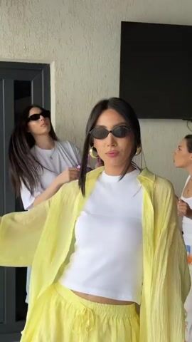 A woman with dark hair and sunglasses is dancing outdoors. She is wearing a white crop top, a yellow open shirt, and yellow shorts. She is also wearing large gold hoop earrings. Another woman with dark hair is visible in the background to the left. The scene appears to be on a patio or balcony.