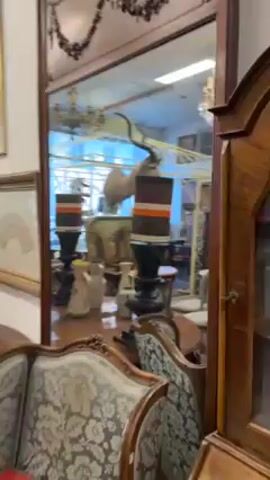 The video presents an interior scene, likely an antique store, displaying a variety of furniture and decorations. A large wooden-framed mirror occupies a significant portion of the view, reflecting the surrounding items, including a chandelier and a woman taking a photo of herself in the mirror. The mirror also reflects parts of the room, showing bookshelves, lamps, and assorted chairs. The camera pans to the right, revealing a taxidermied head of an animal, possibly an antelope or deer, mounted above a decorative cage-like structure. Below the animal head, there is a table with copper pans displayed. The environment is cluttered with various antique items, creating a sense of an eclectic and packed store.