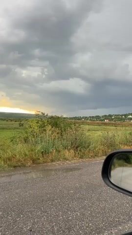 A vehicle's side mirror is visible in the lower right of the frame. The mirror reflects a road and a grey, overcast sky with dark, stormy clouds. The landscape beyond the road features a grassy field with some tall reeds and bushes. In the distance, there are houses and a faint glow of sunlight on the horizon. The road in the foreground appears wet, suggesting recent rain. The overall scene is outdoors and the weather is inclement.
