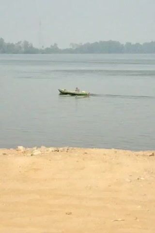 A lone person is in a small green boat on a wide river. The person appears to be operating the boat with an outboard motor, indicated by the wake. The water is calm and grey. The background shows a hazy shoreline with trees and what appears to be a tall radio tower in the distance. The foreground is a sandy bank with some scattered rocks. A small bird flies across the scene in the middle distance. The overall lighting suggests an overcast day.