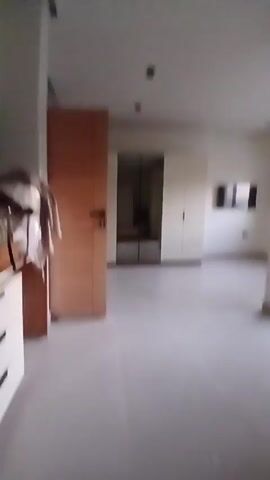 The video shows an empty, spacious room with light-colored walls and flooring. The room appears to be a bedroom or living area. There is a brown wooden door to the left, a mirrored closet in the center, and a white closet to the right. A small, dark-framed window is mounted on the far wall. The video pans to the right revealing a black-framed glass door leading to an outside area.
