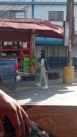 From a moving vehicle, the video shows a woman in a light green outfit walking on a sidewalk.  She passes by a roadside fruit and vegetable stand, with a man standing inside its truck. The video then passes the scene.