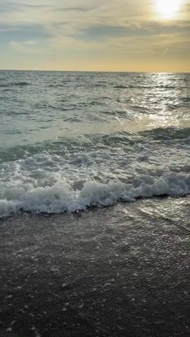 A drone shot hovers above a beach at sunset. The sun, a bright orb, shines through scattered clouds, casting a shimmering reflection on the ocean's surface. Gentle waves roll towards the shore, their white foam lapping at the dark, wet sand. The scene is serene and captures the tranquil beauty of the coast as day transitions to night.