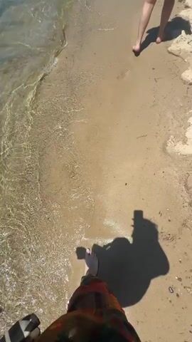 The video captures a first-person perspective of someone walking barefoot on a sandy beach towards the clear water.  Another person, a woman wearing a black bikini top and a tie-dye skirt, walks along the shoreline toward the water. The water is clear enough to see the sandy bottom.  In the distance, other people are visible swimming in the sea.  The sky is clear and blue.  The beach extends along the shoreline to a line of buildings and trees.