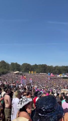 A large, densely packed crowd of people are gathered outdoors, likely at a festival or concert. The day is sunny with a clear blue sky. In the foreground, several individuals are visible from behind, some wearing hats, including a leopard-print cowboy hat and a sequined hat. Further back, the crowd extends as far as the eye can see, with various stages and structures visible in the distance. Colorful flags and banners are scattered throughout the venue. The atmosphere appears festive and energetic, with many people dressed in casual attire suitable for an outdoor event.