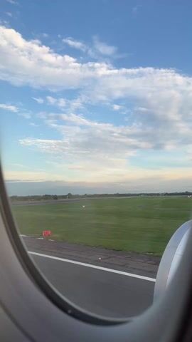 The view from an airplane window shows the runway and surrounding greenery. The sky is blue with scattered clouds. The plane is about to take off or has just landed, as it is moving along the runway. The engine is visible through the window.