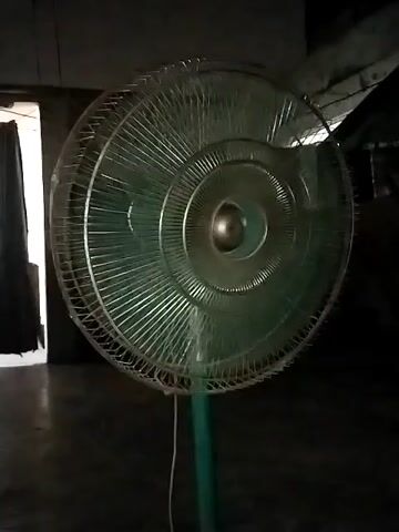 A teal-colored standing fan with a circular metal safety cage is prominently featured in the video. Its blades are actively spinning, indicating it is turned on and operating, producing an audible whirring sound. The fan is positioned on a dark, possibly concrete, floor in a dimly lit indoor environment. To the left in the background, a dark curtain or piece of fabric is visible, partially obscuring what appears to be a doorway or opening. The overall lighting suggests it might be evening, night, or a room with limited natural light. The setting is Dera Chahal, Pakistan. No people or visible text are present in the frame.