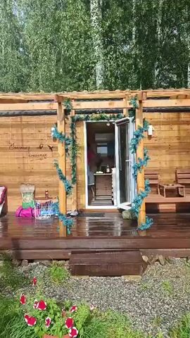 The video pans across a wooden cabin with a covered porch. The porch is wet, suggesting recent rain, and has two wooden chairs and a bench. A green garland is draped over the porch's wooden structure. Inside the cabin, through an open door, a person's legs are visible. The cabin is surrounded by lush green trees, primarily birch and pine trees, with some flowering plants in pots visible in the foreground. A barbecue grill is also visible in the background.