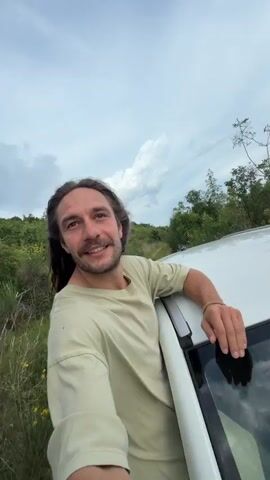 The video begins with a man leaning against a white car, smiling, and giving a "rock on" hand sign. He has long hair and a beard, and is wearing a light green t-shirt. The background features green vegetation with yellow flowers and a cloudy sky. The camera then pans upwards to show wind turbines on a hill covered in greenery under a cloudy sky. The wind turbines are white with long blades that are turning. There are a few trees and yellow flowers in the foreground.