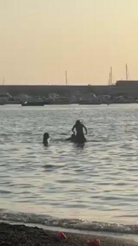 The scene is set at a beach, with the view directed towards the water. Two individuals, one appearing taller, are standing in the water, while another child is swimming nearby. The taller individual seems to be interacting with the other, as they reach out with their arms. In the background, a harbor wall is visible, lined with numerous docked boats. The sky is light, suggesting a late afternoon or early evening setting. The beach itself is visible in the foreground, with dark sand and scattered debris.