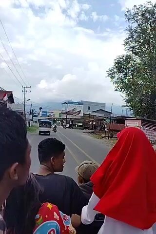 The video shows a group of people riding in the back of an open vehicle. They are looking forward at a street scene. The street is lined with buildings and utility poles. There are vehicles on the road, including a truck and a smaller car. The sky is cloudy with some blue patches. A few people are visible in the foreground, their backs turned to the camera. One person is wearing a red headscarf.