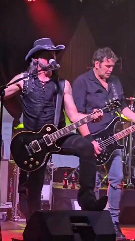 A live music performance takes place on a stage illuminated by red and green lights. A man in a cowboy hat and a black vest with straps plays a black electric guitar. To his right, another man in a dark shirt and ripped jeans plays a black bass guitar. In the background, a drummer is visible, seated behind a drum set with a logo featuring a winged scorpion. A lead singer with long dark hair, wearing a black shirt and pants adorned with skull patterns, is on the far right, holding a microphone and singing. The stage is equipped with various musical instruments, microphones, amplifiers, and stage lighting.