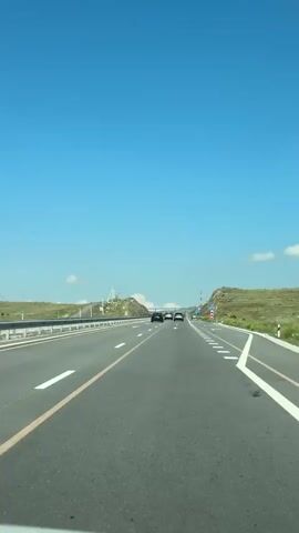 The video shows a forward-facing perspective from inside a vehicle traveling on a highway. The road is paved, with distinct white lane markings and a yellow dividing line. Several cars are visible ahead, traveling in the same direction. The landscape on either side of the highway consists of rolling green hills under a clear blue sky. There are signs and guardrails along the side of the road.