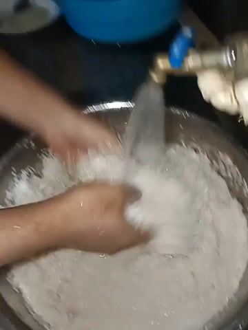 The video shows a close-up of a person's hands washing rice in a metal bowl under a running faucet. The rice appears to be white and dry initially, but becomes submerged in cloudy water as it is rinsed. The person's hands are actively moving the rice around in the bowl, agitating it to remove any impurities. The faucet is a standard metal fixture, and the water stream is strong and clear. The environment appears to be a kitchen sink area, with some out-of-focus background elements suggesting a domestic setting. The lighting is adequate for viewing the activity.