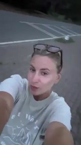 A blonde woman with sunglasses on her head films herself in selfie mode. She is wearing a white t-shirt. She sticks her tongue out at the camera and then resumes filming as she is moving. In the background is water and a paved surface. The light is fading.