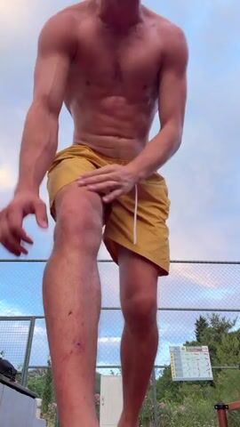 This is a video of a shirtless man in yellow shorts performing a backflip outdoors. The man has long hair and a beard, and appears to be fit. He is seen in a standing position, then jumps and flips, landing back on his feet. The backdrop is a cloudy sky, with a fence and some trees visible. There is a sign in the background with what looks like exercise instructions, featuring stick figures demonstrating various physical activities. The lighting suggests it is late afternoon or early evening, with the sky showing hints of warm colors from the setting sun. The location is identified as Petrovac na Moru, Montenegro. The man's expression at the beginning and end of the flip suggests focus and accomplishment.