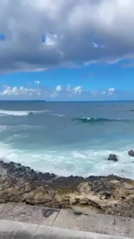 The video shows a coastal scene with ocean waves crashing against a rocky shoreline. The water is a mix of light blue and turquoise. Further out in the water are a few dark shapes, possibly people surfing. The sky is blue with fluffy white clouds. In the foreground is a rocky ledge with some green vegetation and a grey concrete barrier.