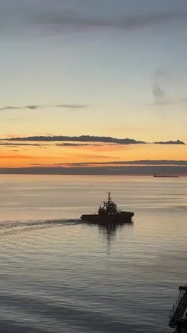 A tugboat is moving through calm water at sunset. The sky is a gradient of blue and orange, with some clouds. The water is reflecting the colors of the sky. The tugboat is silhouetted against the sunset, and its wake is visible behind it.