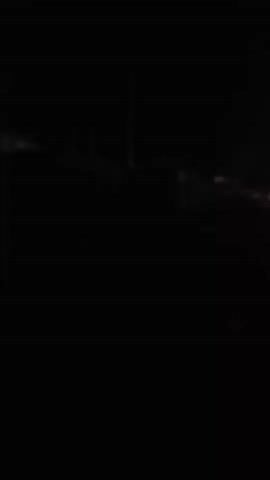 The video is very dark, with most of the scene obscured by shadows. In the first few frames, there appears to be some structure or building in the background with vertical elements, possibly poles or pillars. A faint light source is visible in the distance. As the video progresses, the scene remains largely dark, with occasional flashes of light or movement that are difficult to discern. In the final frame, there is a bright light source, possibly a streetlamp or a powerful flashlight, illuminating a small area of what appears to be foliage or vegetation. The rest of the scene is still dark and indistinct. There are no visible people or clear actions taking place. The environment appears to be outdoors, possibly at night, given the darkness and artificial lighting.