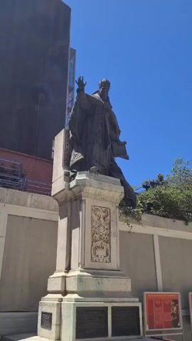 The video shows a low-angle shot of a bronze statue of a robed figure, possibly a religious leader, on a stone pedestal. The statue is located outdoors, with a bright blue sky and some greenery visible in the background. The pedestal has ornate carvings and plaques at its base. To the right of the statue, part of a building with architectural details and some banners is visible. Several framed exhibits or posters are displayed on the ground in front of the building. The overall impression is of a historical or religious site on a sunny day.