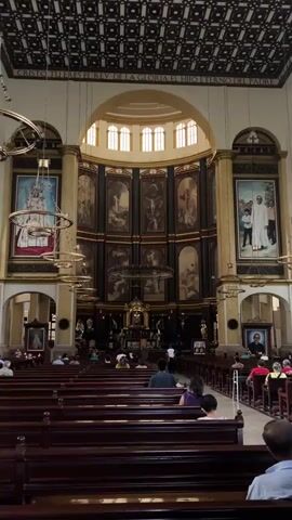The interior of a church is shown in the video. Rows of brown wooden pews stretch from the camera towards the front of the church, and people are seated in them. A high altar is visible at the front, with large paintings displayed behind it. The walls are painted in shades of cream and green. Large chandeliers hang from the high, ornate ceiling. A man walks from right to left between pews.