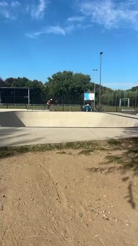 The video shows a skateboard park on a sunny day. In the background, a person is skateboarding in the bowl. They are wearing shorts and a hat. To the right of the skateboarder, a person is sitting on the edge of the skate park, looking at their phone. They are wearing a black shirt and black shorts. The skate park is made of concrete and surrounded by grass and trees. There is a fence in the background.