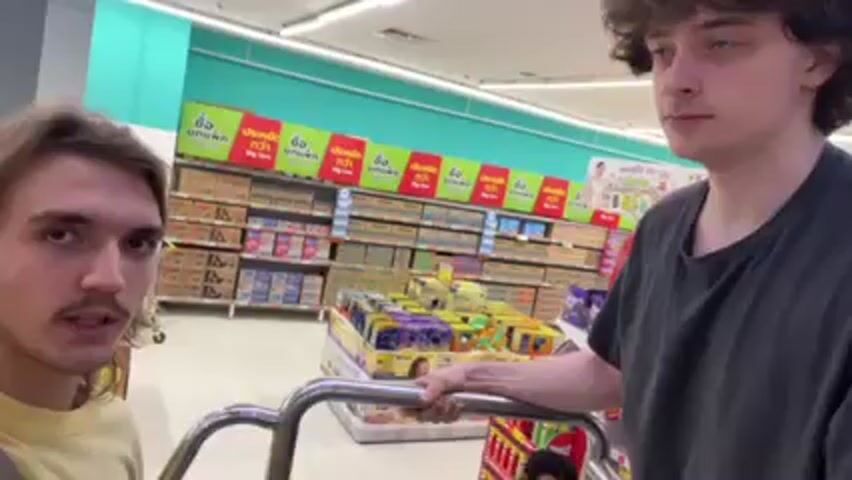 The video shows two young men at a supermarket, standing by a chrome-plated shopping cart. The person filming is wearing a yellow T-shirt and a shoulder bag. He has long hair and a mustache. The second man is wearing a dark T-shirt and is standing behind the shopping cart. They appear to be discussing something. Behind them are shelves stocked with various products. The background has colorful, repeating text.