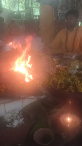The video shows a religious ceremony. A man in a yellow robe is sitting near a fire, holding a ladle. There are flowers and other offerings around the fire. A candle is also burning. The man appears to be chanting or praying. The ceremony is taking place indoors, likely in a temple or home.