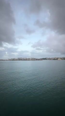 The video shows a body of water with a city skyline in the distance under a cloudy sky. The camera appears to be moving, as evidenced by the slight shift in perspective of the water and the skyline. The water is a deep teal color and appears choppy with small waves. The skyline consists of buildings of varying heights, some of which are brightly colored. The sky is filled with large, gray clouds, with patches of lighter sky peeking through. The overall mood of the video is somber and atmospheric.