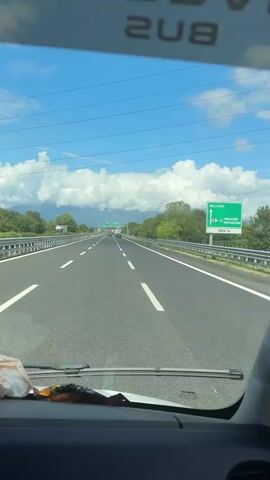 A sped-up video shows a view from a vehicle traveling on a highway. The road has multiple lanes and is bordered by a metal guardrail and trees. Overhead signs indicate directions to "BELLUNO" and "CONEGLIANO PORTOGRUARO". The sky is blue with scattered clouds, and mountains are visible in the distance. Other vehicles, including cars and a truck, are also on the highway. The video appears to be captured during daylight hours, likely in the summer.