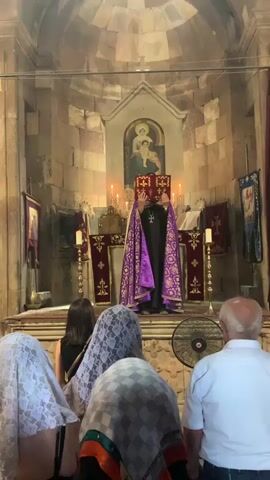 A priest in purple vestments holds an open book above his head in front of an altar. Candles are lit on either side of the altar, and a painting of the Virgin Mary and child is visible behind the priest. Several people, mostly women wearing head coverings, are kneeling in the foreground, facing the altar. The scene appears to be inside a stone church, with a beam of light shining down from a ceiling opening.