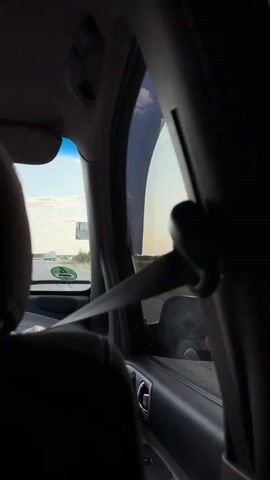 The video is shot from inside a moving vehicle, looking out the passenger side window. The camera pans to the right, showing a rural landscape. In the foreground, the interior of the car is visible, including the seatbelt and the car's pillar. Outside the window, there is a road, a field of what appears to be sunflowers, some trees, and a bright sky with scattered clouds. The sun is visible in the upper right corner of the frame, suggesting it is either early morning or late afternoon. The video captures the feeling of a scenic drive through the countryside.