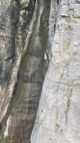 The camera focuses on a tall rock formation with a thin waterfall flowing down its side. The rock is gray and layered, with some patches of green moss clinging to it. The camera pans up the rock face, showing the texture and height. There is also some indistinct chatter in the background.