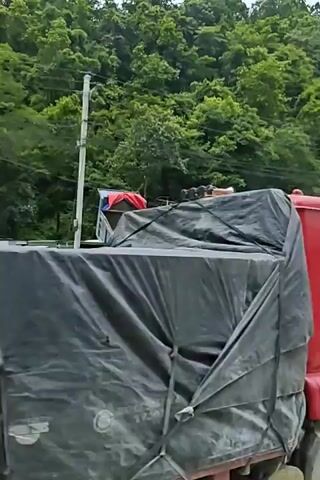 The video shows a dusty, rural road with several trucks parked. In the foreground, a blue-bed truck is visible. Behind it, a red truck's cab is tilted at an extreme angle, as if being unloaded or repaired. Further back, a yellow loader and another truck are parked. The scene is bordered by a steep, tree-covered hillside. Buildings, some appearing under construction, are visible along the road. The sky is cloudy.