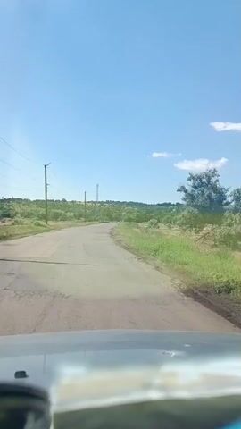 This video shows a view from inside a vehicle driving down a cracked asphalt road through a rural landscape in Artsyz, Ukraine. The sky is a clear, bright blue with a few scattered white clouds, suggesting it is daytime and the weather is fair. The road is lined with dry grass and scattered trees. In the distance, rolling hills covered in vegetation are visible, along with some power lines and a tall transmission tower. The overall mood is one of travel and open space. There is no visible text in the media.