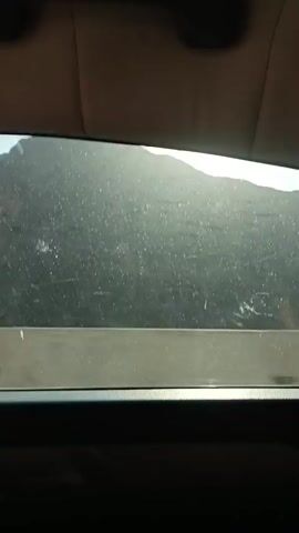 The video shows a view through a car window. The sun is shining brightly, creating lens flare across the image. In the distance, a mountain range is visible, with some trees or vegetation on its slopes. There are power lines visible on the mountain. The interior of the car is visible at the bottom of the frame, with a speaker grill. The video appears to be recorded while the car is in motion.