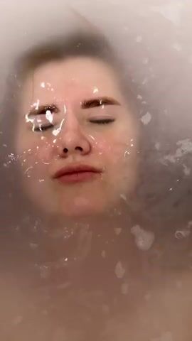 A close-up video shows a woman submerged in a bathtub, looking directly at the camera. Initially, her eyes are closed, and her face is mostly underwater, with some bubbles surrounding her. She then slowly lifts her head out of the milky, bubbly water, opening her eyes and revealing a slight smile. Her face is wet, and her dark hair is slicked back. She has light skin, visible freckles, and dark eyebrows. The setting is indoors, in what appears to be a bathroom. The woman's expression is playful and content. No text is visible in the video.