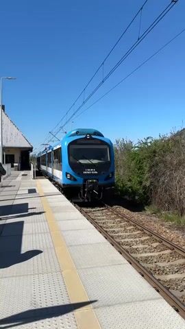 The video captures a blue, black, and white train departing from a station platform on a clear, sunny day in Ustroń, Poland. The train, identified as "EN57AKŚ-1533ka" on its front and "EN57AKŚ-1533ks" on its side, also displays the numbers "2 120 811-6" and "94 51 2 120 812-4 PL-KŚL". The "KOLEJE ŚLĄSKIE" logo is visible on its side, along with a bicycle symbol, "UWAGA!" warning signs, and the number "2" prominently displayed near its yellow doors.

The camera is positioned on the textured platform, which has a yellow tactile strip along its edge, facing the train as it moves from left to right. Overhead power lines stretch across the clear blue sky. To the left, a portion of a building with a light-colored roof can be seen, while green bushes line the right side of the tracks. Strong shadows on the platform indicate bright sunlight.