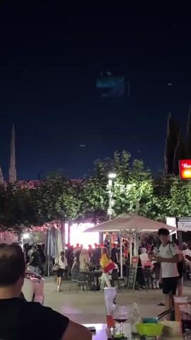 A vertical video shows a crowd of people gathered at night in an outdoor area.  There is a large screen displaying a vibrant image, possibly a sports event or performance, with bright colors and figures.  Several trees with green foliage are visible, some illuminated by spotlights.  People are standing and sitting at tables, some with umbrellas, suggesting an outdoor dining or viewing area.  One person in the foreground is wearing a Spanish flag draped over their shoulders. The overall atmosphere appears festive and lively.
