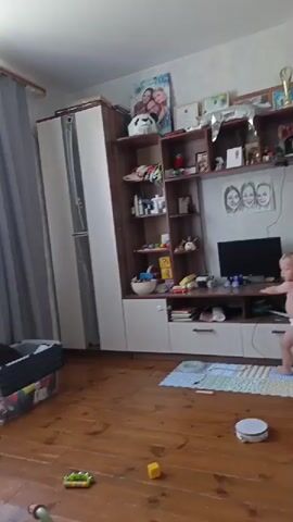 The video begins by showing a toddler wearing a diaper standing in a room in front of a large entertainment unit. The toddler is facing the entertainment unit and appears to be reaching for something. The room has wooden floors and a large window with curtains. There are several objects visible on the entertainment unit, including pictures, trophies, and various decorative items. Toys are scattered on the floor. The scene then cuts to a different part of the room, where the toddler is now wearing pajamas and is on a couch, leaning forward as if looking under it. The window is still visible in the background. The video ends with a wider shot of the room, showing the couch, the entertainment unit, and the window.