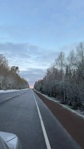 The video shows a car driving on a paved road through a wintry landscape. The road is grey with a white dividing line, and there's a narrow red shoulder on the right. Trees line both sides of the road, their branches lightly covered with snow. The sky is a mix of blue and grey clouds, with a hint of orange on the horizon in the distance. The overall scene is peaceful and serene.