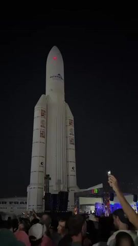 At night, a crowd of people have gathered in front of a large, white model of a rocket. The rocket is labeled "ARIANESPACE" and has flags and the letters "ESA" and "CNES" on its sides. Stage lighting casts blue hues on the rocket, the crowd, and the stage in front of the crowd. The video shows the rocket from various angles, illuminated by the stage lighting. Many people are holding up their phones to capture the scene. An airplane is visible behind the crowd.