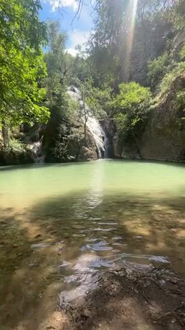 A serene natural scene unfolds featuring a secluded waterfall cascading into a shallow, clear turquoise pool. Lush green foliage drapes the rocky cliffs surrounding the pool, while sunlight filters through the trees, casting dappled shadows on the water's surface. The camera remains stationary, focusing on the tranquil beauty of the waterfall and its pristine surroundings.