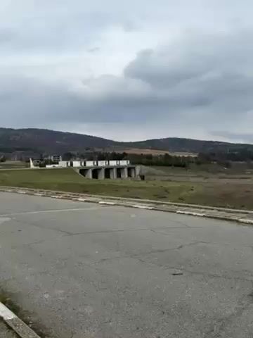 The video shows a concrete dam structure on a grassy hillside under a cloudy sky.  In the background, there is a forest and distant hills. The camera pans slightly to the left, revealing more of the landscape and then to the right showing more of the dam.  There is a cracked asphalt road in the foreground.