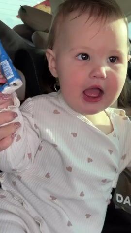 A baby with light brown hair, wearing a white long-sleeved shirt adorned with small brown hearts, is seated in a car. She holds a white and blue object in her left hand and a white and pink object in her right. Throughout the video, the baby displays a joyful demeanor, smiling broadly, looking around, and opening her mouth as if laughing. Behind her, partially visible, is a woman with dark hair, who also appears to be looking forward or around the car. Part of a bag with the text "DA BLY" is visible next to the baby. The interior of the car is visible in the background, suggesting they are on a journey.