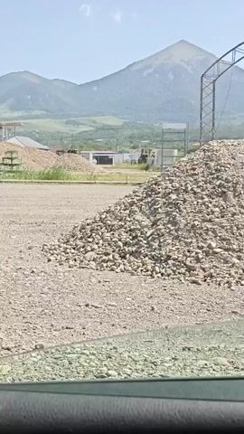 A gravel lot is in the foreground. A large pile of rocks is on the right side of the frame. In the background are more piles of rocks, buildings, a crane, and mountains. The sky is blue with a few clouds. A car window is visible at the bottom of the frame.