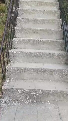 The video shows a first-person perspective of someone walking up a long, steep, concrete outdoor staircase. The stairs have metal railings on both sides. The surrounding environment is rocky and appears to be a natural landscape with some vegetation. The sky is visible and blue. The video progresses upwards as the person ascends the stairs, revealing more of the rugged terrain and the continuing staircase.