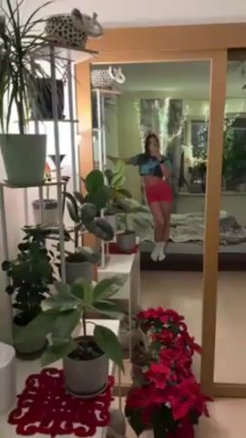 A young woman is posing for a selfie in front of a large, framed mirror. She is wearing a tie-dye crop top, bright pink shorts, and white socks. Her hair is down. The room reflected in the mirror shows a bed, curtains with string lights, and various plants. The area in front of the mirror has a tall plant stand with multiple plants, a small white table, and a cluster of bright red poinsettia plants. The mirror has a gold-colored frame.