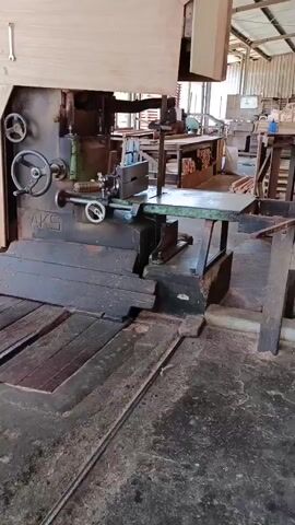 The video begins by showing a large industrial saw, specifically a band saw, in a workshop. The saw is made of dark metal and has a large wheel for adjusting the blade tension. There are some wooden planks on the floor in front of the saw, and sawdust is visible. The camera then pans to the left, revealing more of the workshop environment. We see stacks of lumber and wood, as well as some metal shelving units. The background shows an outdoor area with trees and buildings, suggesting the workshop is located in an industrial yard or lumber mill. The overall environment appears to be a functional workspace, possibly a carpentry shop or a place where wood is processed. There are no people visible in the footage. The lighting suggests it is daytime, with sunlight filtering in from the open-air sections of the workshop.
